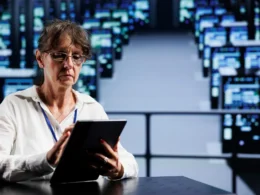 Woman with glasses using a tablet while seated at a table in a data center environment.